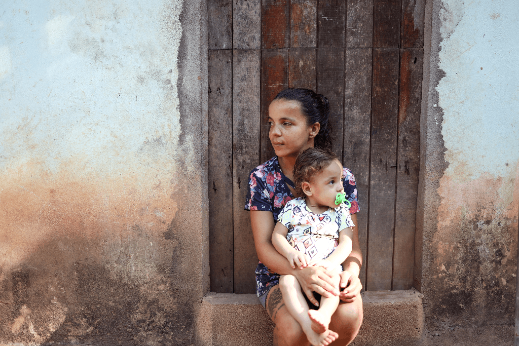 Carla, wearing a black shirt with a floral print, is sitting in front of her neighbour's door holding her daughter, Clara.