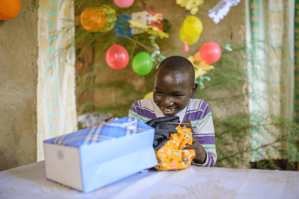 Elia is wearing a purple and white striped shirt. He is sitting at a table and is unwrapping his Christmas gift.