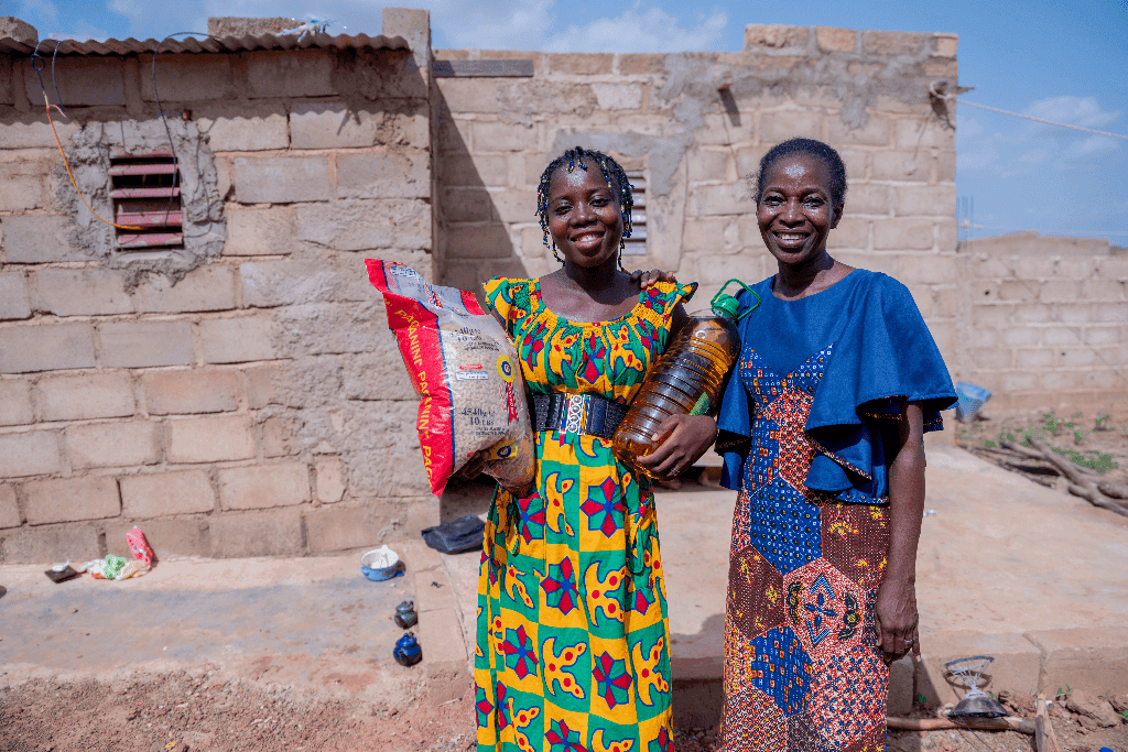 Wennefangdé is wearing a yellow, red, and green patterned dress. She is standing outside and is holding a bag of pasta and a bottle of oil she received from Compassion. Next to her is Ahoua, Centre Director.