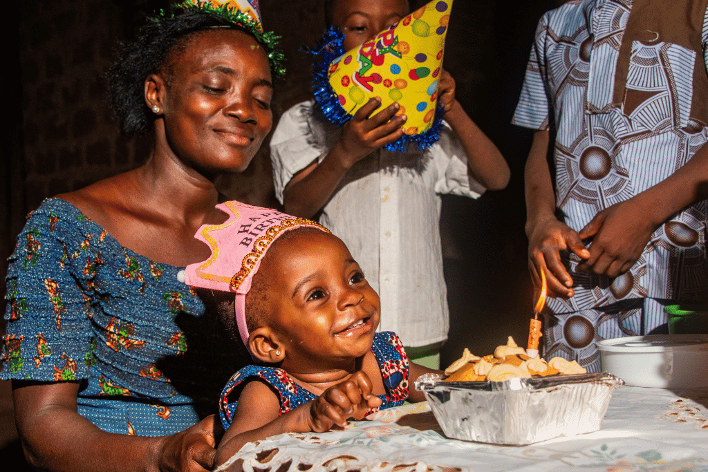 Emmanuella, in a blue dress and pink birthday crown, is smiling as she sits on her aunt Mawulawoe's lap