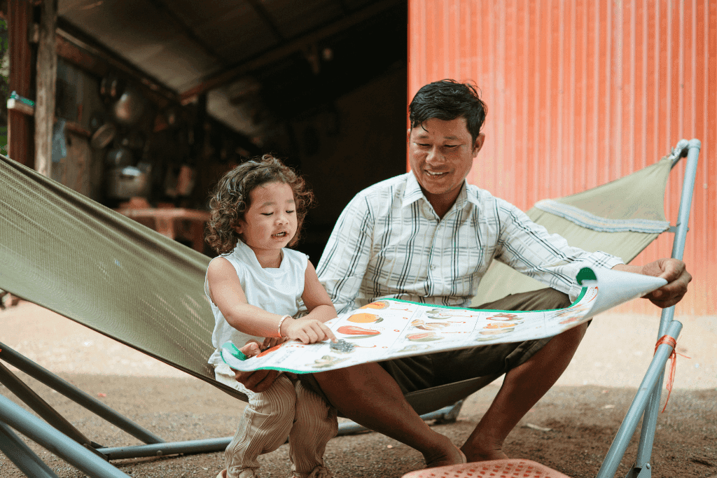 Monika sits on the green hammock with the poster spread out over her and her dad's lap as they look down at to a section of the poster she is pointing at. Her dad is smiling as he listens to her.