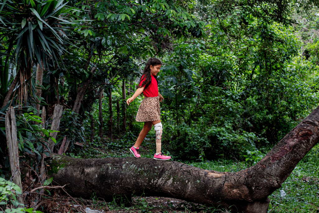 Connie walks on top of a large tree branch. She is surrounded by green trees.
